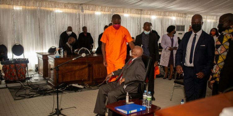 Ex-president Guebuza, seated, talks to his son Ndambi Guebuza, wearing an orange prison uniform. The younger Guebuza is a defendant in the long-awaited trial, which is unfolding in a marquee in a high-security jail | AFP
