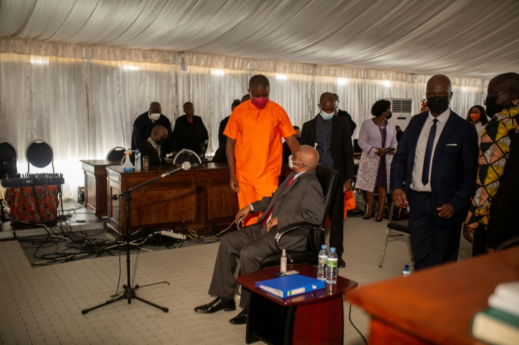 Ex-president Guebuza, seated, talks to his son Ndambi Guebuza, wearing an orange prison uniform. The younger Guebuza is a defendant in the long-awaited trial, which is unfolding in a marquee in a high-security jail | AFP
